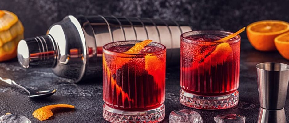 Ръка държи чаша червен коктейл с кубчета лед и гранули тартюфи отгоре, опряна върху тъмна повърхност.